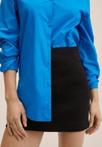 Chemise boutonnée en tissu bleu vif associée à une mini-jupe noire texturée, mettant en valeur des couleurs et des matériaux contrastants.