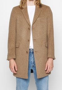 Henkilö, joka on pukeutunut nappilistalla varustettuun kamelinruskeaan villapaitaan valkoisen paidan ja vaalean sinisten farkkujen päälle, seisoo yksinkertaista taustaa vasten.