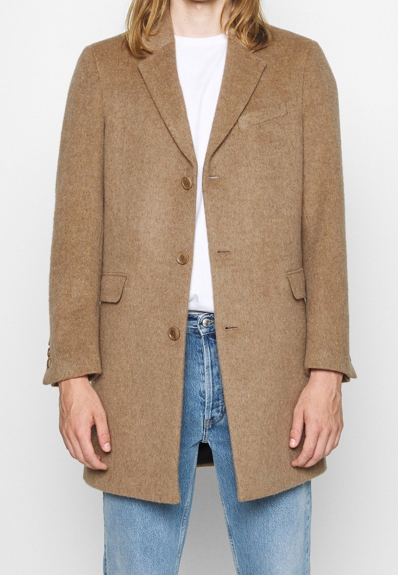 Henkilö, joka on pukeutunut nappilistalla varustettuun kamelinruskeaan villapaitaan valkoisen paidan ja vaalean sinisten farkkujen päälle, seisoo yksinkertaista taustaa vasten.