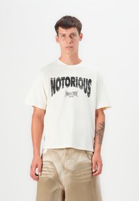 Cream-colored short-sleeve t-shirt featuring bold black “NOTORIOUS” graphic text and "World Tour 1990" underneath. Soft cotton material.
