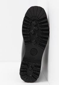 Semelle d'une botte noire avec des crampons profonds comprenant des motifs en forme de croix et linéaires, inclinée contre un mur blanc à angle.