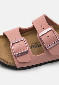 Világos rózsaszín Birkenstock szandál, amelyen két állítható pánt található fém csatokkal, parafa talp és texturált gumitalp.