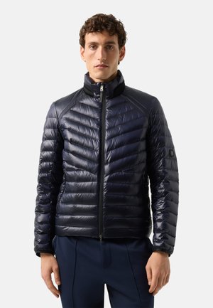 Mann trägt eine dunkelmarineblaue gesteppte Daunenjacke mit Reißverschluss, steht vor einem schlichten hellen Hintergrund.