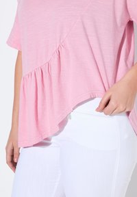 Světle r�ůžový top s krátkým rukávem, který má nařasený lem a asymetrický design, v kombinaci s bílými kalhotami. Viditelná je měkká textura látky.