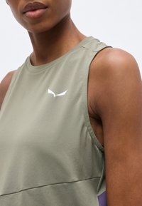 Κοντινό πλάνο ενός ατόμου που φορά ένα αμάνικο ελαφρώς ελιάς τοπ με ένα μικρό λευκό λογότυπο στο στήθος, μπροστά από απλό φωτεινό φόντο.