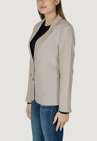 Ανοιχτό μπεζ σακάκι με έναν μόνο κουμπί, αιχμηρό πέλμα και δύο μπροστινές τσέπες. Συνδυάζεται με μαύρο πουκάμισο και μπλε τζιν. Λείος υφασμάτινος. 