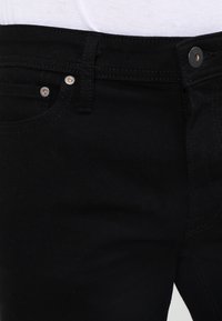 Svarta denimjeans med en slät yta, med kopparfärgade knappdetaljer och en standard femfickdesign.