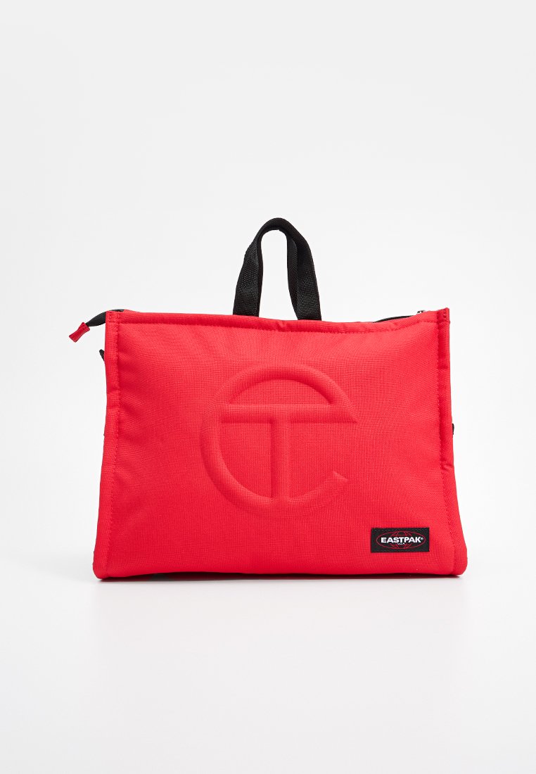 Sac en tissu rouge au design minimaliste, avec un logo en relief et des poignées noires. Fermeture éclair sur le bord supérieur.