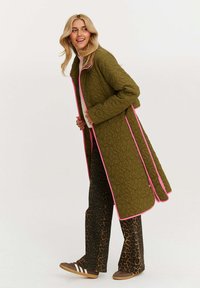 Dlhý prešívaný kabát olivovo zelenej farby s ružovým lemovaním, skombinovaný s hnedými rozšírenými nohavicami s leopardím vzorom a hnedými teniskami s bielymi pruhmi.