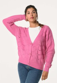Roze cardigan met een gestructureerd ontwerp dat golfpatronen bevat, geribde mouwen en knopen aan de voorkant. Draag het over een witte top, gecombineerd met blauwe jeans.