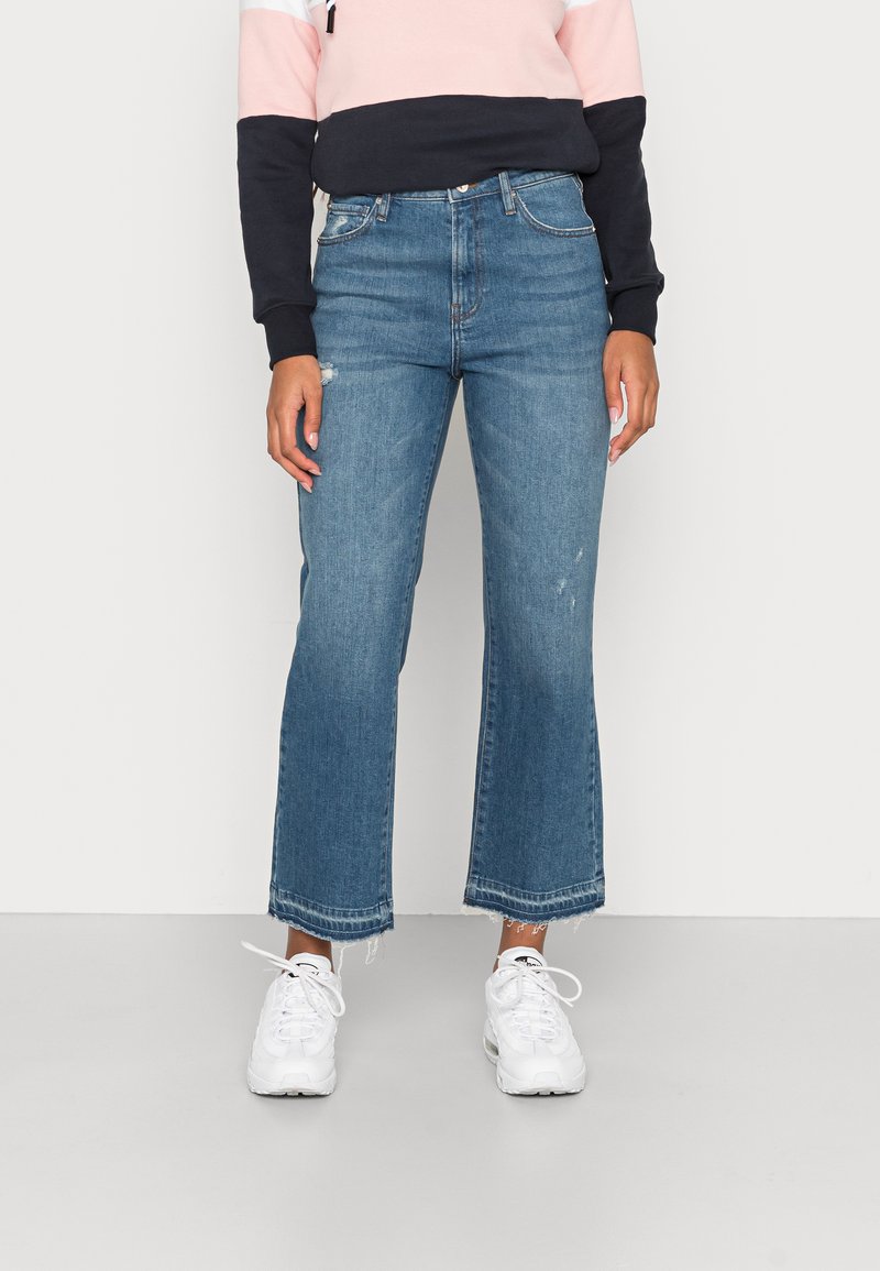 Jeans cortados azul claro com a bainha ligeiramente desfiada, apresentando um corte relaxado e um ligeiro desgaste, combinados com sapatilhas brancas.