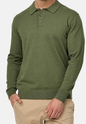 Man draagt een olijfgroen poloshirt met lange mouwen en geribbelde manchetten, gecombineerd met beige broek. Zijn handen rusten vlakbij de zakken.