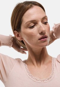 Femme aux cheveux châtain clair portant un haut côtelé rose clair avec un bord en dentelle, levant les bras derrière la tête sur un fond blanc.