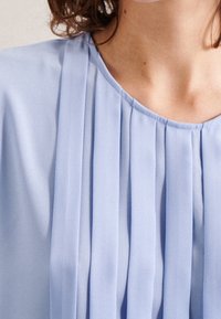 Blusa azzurro chiaro con design a pieghe, stoffa morbida, scollatura rotonda e leggera lucentezza. La superficie testurizzata aggiunge profondità.