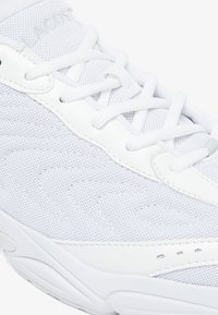Chaussure de sport blanche avec un dessus en maille et des détails texturés, des accents synthétiques et des lacets plats blancs. Semelle en caoutchouc avec des éléments de design subtils.