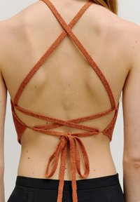 Offene, cropped Bluse mit gekreuzten, strukturierten orangen Trägern; am Rücken gebunden. Glatte Hauttextur und einfacher schwarzer Bund sichtbar.