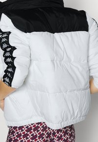 Czarna i biała kurtka puchowa z pikowanym wzorem, elastycznymi mankietami oraz logo po bokach rękawów. Teksturowana tkanina.
