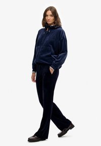 Vrouw in een marineblauw velours hoodie en bijpassende broek, staand met één hand in de zak en donkere chocoladebruine schoenen op een witte achtergrond.