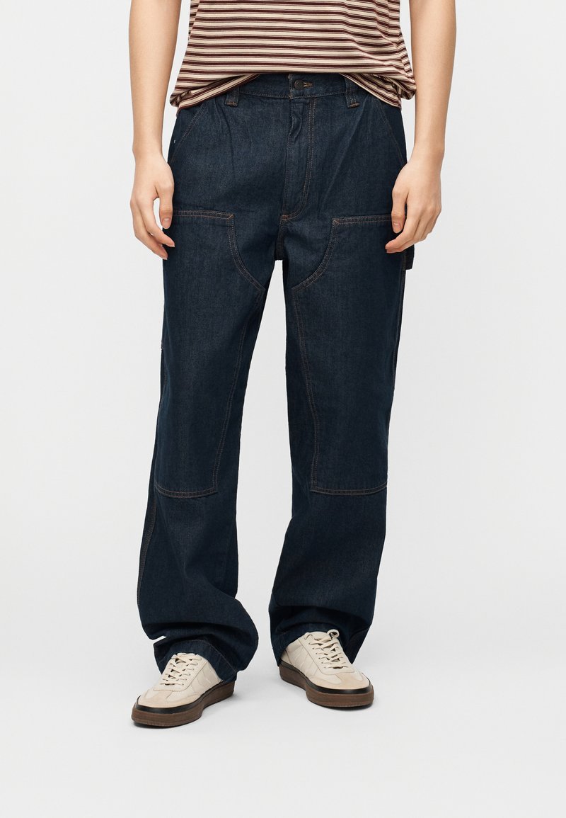 Άτομο που φοράει χαλαρά σκουρομπλε τζιν παντελόνια με μεγάλες μπροστινές τσέπες και λευκά αθλητικά παπούτσια, στέκεται σε απλό λευκό φόντο.