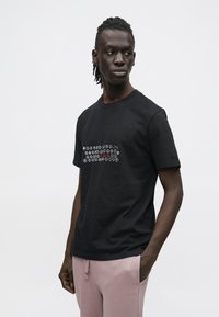 Juoda medvilninė marškinėlių su grafiniu tekstu balta ir raudona spalvomis ant krūtinės. Trumpomis rankovėmis ir klasikine apvaliomis iškirpte.