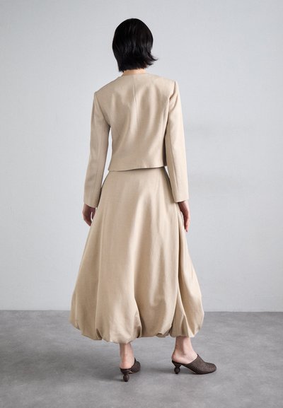 Veste en lin beige et jupe volumineuse avec un ourlet arrondi. Mules tissées sombres avec un design de talon unique. Fond minimaliste.
