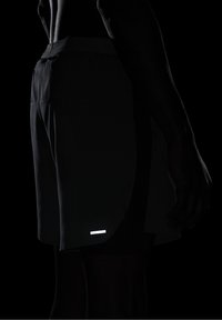 Personne portant un short noir avec une petite bande réfléchissante blanche près de l'ourlet, vue de profil sur un fond sombre.