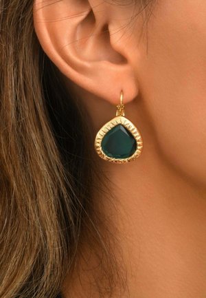 Gros plan d'une oreille portant une boucle d'oreille en or avec une grande pierre précieuse triangulaire vert foncé, suspendue à un petit anneau, avec des cheveux bruns visibles.