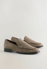 Mocassins pour hommes en suède brun avec des talons bas noirs et des détails de couture minimalistes, présentés côte à côte sur un fond clair.