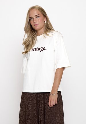 Blonde Frau trägt ein weißes T-Shirt mit dem Text "vintage." und einen braunen gemusterten Rock, steht vor einem schlichten weißen Hintergrund.