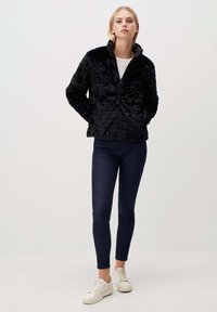 Zwarte nepbontjas met een gequilt patroon, hoge kraag en ritssluiting. Draagt met donker slim-fit jeans en witte sneakers.