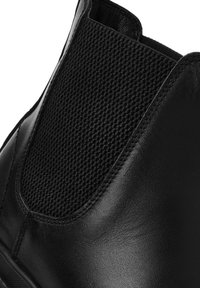 Primer plano de una bota Chelsea de cuero negro con panel lateral elástico texturizado y costuras detalladas.