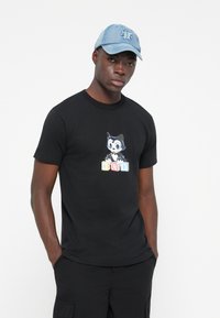 Black cotton t-shirt featuring a cat graphic in blue and white holding colorful blocks reading "HUF." Paired with a light denim cap.