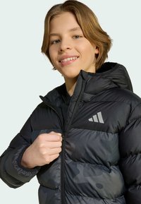 Doudoune noire avec capuche, présentant un motif camouflage subtil, une texture brillante et un logo gris sur le côté gauche de la poitrine. Fermeture zippée.
