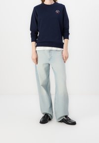 Navy sweatshirt with round neckline and embroidered detail on chest, paired with light blue wide-leg jeans and black loafers.