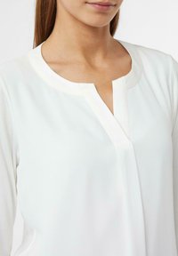 Frau trägt eine weiße Langarmbluse mit Rundhalsausschnitt und einem kleinen V-förmigen Ausschnitt in der Mitte.