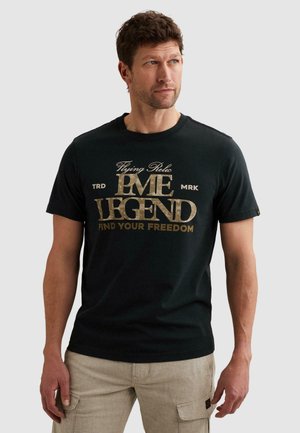 Muž v čiernom tričku PME Legend so zlatým textom "Flying Relic PME Legend Find Your Freedom" a béžových cargo nohaviciach, pozerá vpravo