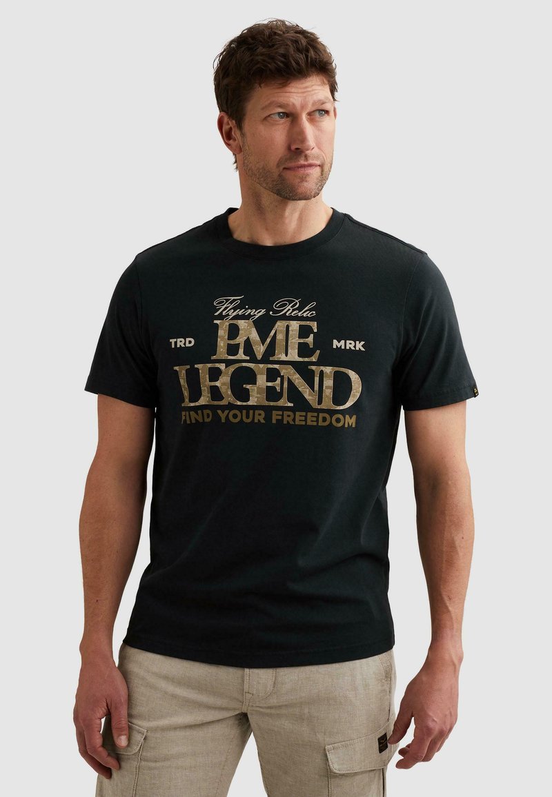 Homme portant un t-shirt noir PME Legend avec texte doré "Flying Relic PME Legend Find Your Freedom" et un pantalon cargo beige, regardant vers la droite