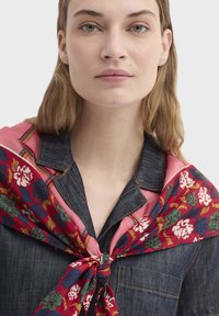 Écharpe en soie avec un motif floral vibrant en rouge, vert et bleu, nouée autour du cou sur une chemise en denim foncé avec des coutures contrastées.