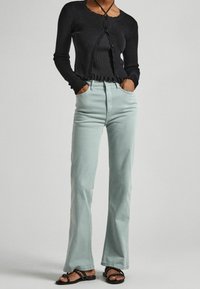 Black long-sleeve cardigan over a black ribbed top, paired with light green flared jeans and black sandals with studded accents.