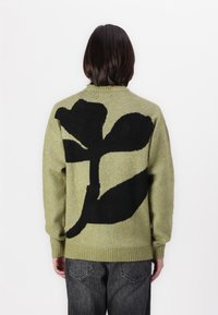 Sweter dziergany w jasnym oliwkowym kolorze z dużym czarnym kwiatowym wzorem na plecach. Teksturowana tkanina z prążkowanymi mankietami i obrębem.
