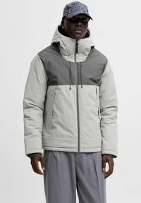 Grau und hellgraue Steppjacke mit Kapuze, ausgestattet mit einem Reißverschluss vorne und Kordeln. Kombiniert mit lässigen grauen Hosen und einer Mütze.