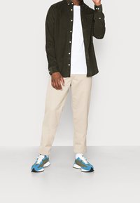 Outfit maschile composto da una camicia a maniche lunghe in velluto a coste verde scuro con bottoni, t-shirt bianca, pantaloni beige e sneakers blu/verdi con dettagli neri.