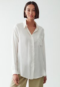Camicia button-up bianca in seta con colletto, maniche lunghe e taschino frontale. Texture liscia e vestibilità rilassata, con orlo curvo.