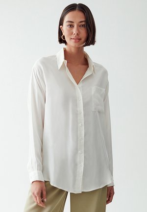 Camicia button-up bianca in seta con colletto, maniche lunghe e taschino frontale. Texture liscia e vestibilità rilassata, con orlo curvo.