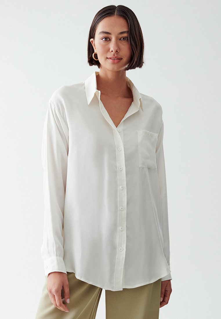Camicia button-up bianca in seta con colletto, maniche lunghe e taschino frontale. Texture liscia e vestibilità rilassata, con orlo curvo.
