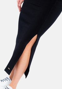 Czarna, żeberkowana spódnica midi z bocznym rozcięciem, prezentująca gładką fakturę i złoty akcent na końcu. Zestawiona z białymi sneakersami.