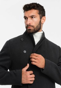 Abrigo de lana negro con un cuello alto, que cuenta con tres botones. El modelo está ajustando el cuello sobre un suéter de cuello alto blanco.
