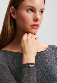 Schwarzes glitzerndes Oberteil mit Schiffsausschnitt und langen Ärmeln. Besitzt einen silbernen Akzent am Bündchen. Glatte Textur und taillierter Schnitt.
