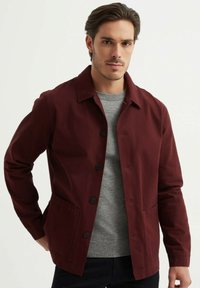 Veste bordeaux en tissu lisse, dotée d'un col classique, de boutons sur le devant et de deux poches latérales. Portée sur un pull gris.