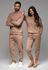 Matchande beige sweatshirt och joggers i mjukt material, med ribbade mudd och subtil branding. Den manliga modellen bär vita sneakers, medan den kvinnliga modellen har beige tofflor.